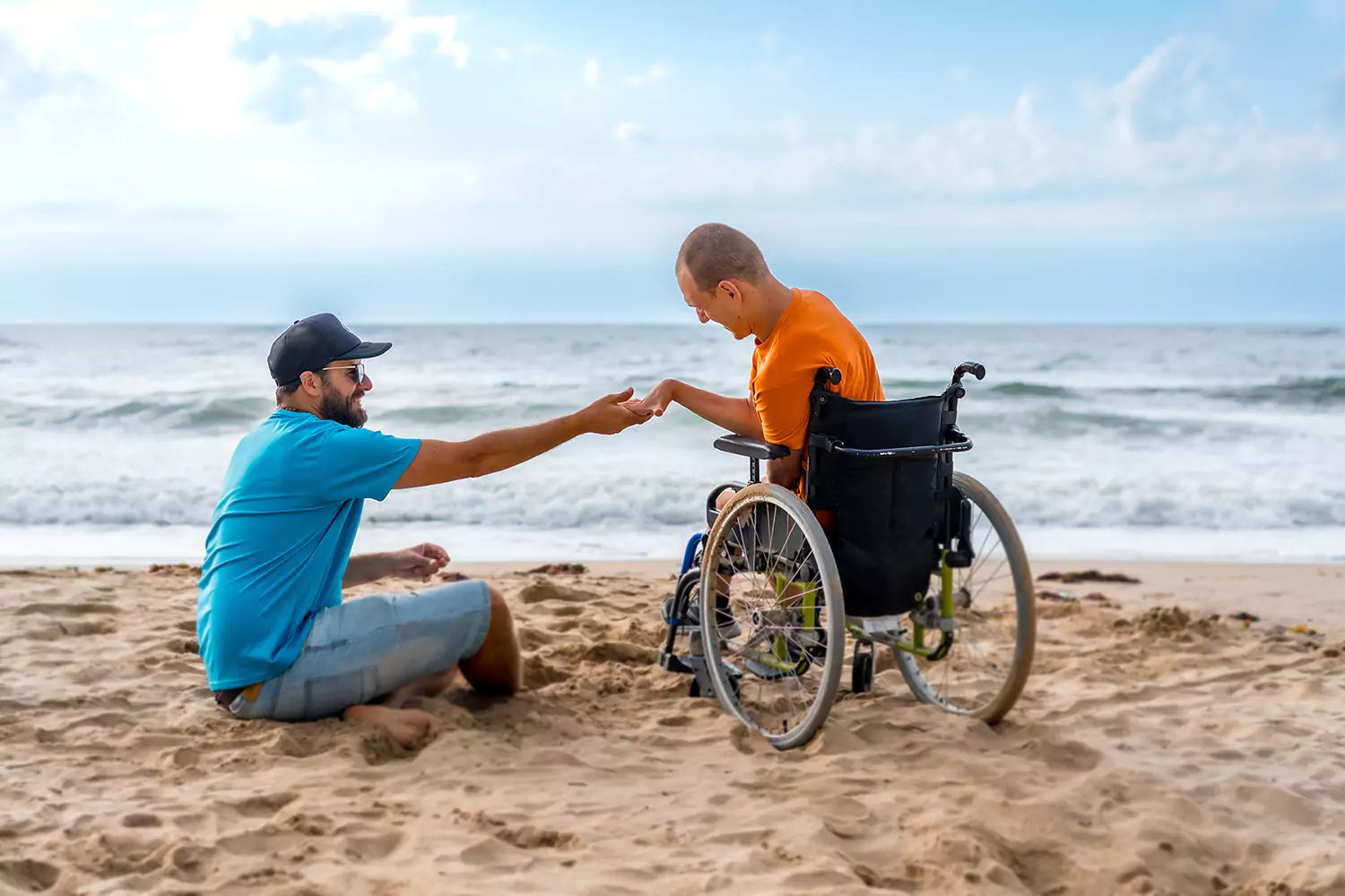 A disabled man sitting in his wheelchair and his support worker in the beach holding hands